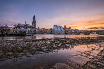 Fototapeta premium Sonnenuntergang während Hochwasser des Flusses Donau im Winter 2021 in Regensburg mit Blick auf den Dom die Altstadt und überschwemmte Promenade und der steinerne Brücke, Deutschland