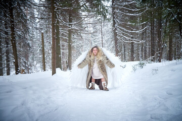 A middle-aged woman in a large warm fur coat and white angel wings in a winter forest with snow and snowdrifts. Fairy angel in a cold forest in Russia before Christmas