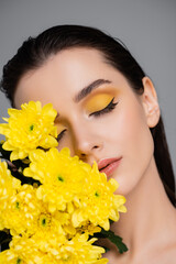 young brunette woman with makeup near blooming yellow flowers isolated on grey