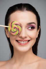 happy young woman with pink eye shadows covering eye with lollipop isolated on grey