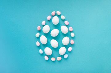 White chicken eggs and small colored eggs are laid out in the form of a large egg in the center on a blue background. Easter creative design, greeting card. Top view, flat lay, copy space.