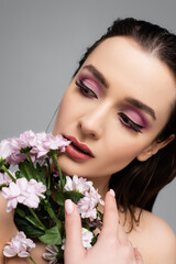 young woman with pink eye shadows looking away near flowers isolated on grey