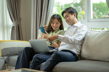Asian couple smiling and looking at each other happily chatting. Are shopping online at home, using internet banking services.