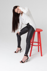 A beautiful girl of 25, European appearance, with dark long hair, in a white shirt and black trousers, sits on a bar stool in a light studio and looks pensive. Generation Y