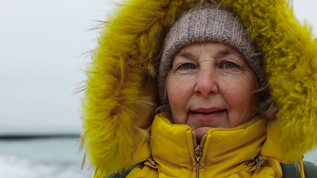 An elderly tourist woman in warm bright clothes with a backpack walks on the seafront by the sea in winter weather. Senior woman tourist looks at camera