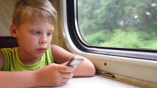 Closeup view video portrait of attentive little white boy traveling by train and using mobile phone while sitting at table in moving wagon of train. Green summer trees passing by in background