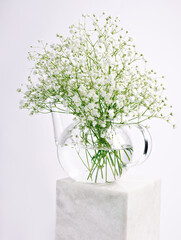 Glass vase with bouquet of fresh field flowers sitting on the bright marble cube. Minimal spring bloom concept. Neatly arranged visual trend layout.