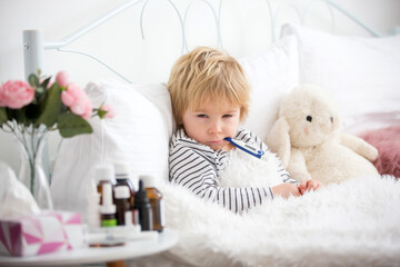 Sick little blond toddler boy, lying in bed with fever, hugging little fluffy toy, medicine next to him