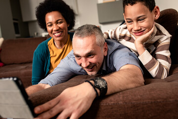 Smiling mixed race family enjoying time at home laying on sofa in living room making selfie with tablet.