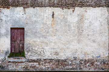 Old wall with window. Aged wall painted in white and with a red wooden door that seems abandoned.