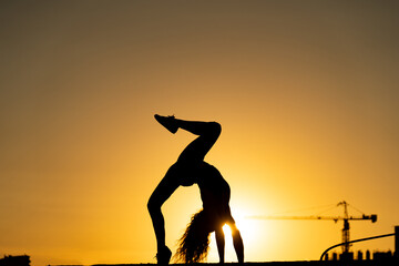 Female gymnast showing her flexibility and split during sunset on orange sky background with crane and construction. Concept of freedom and happiness