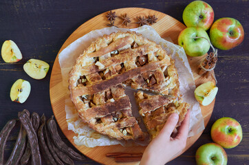 Hand takes a piece of traditional apple pie with spices. Apple tart  decorated with sliced fresh apples, carob pods and cinnamon sticks on the dark bakground. Top view