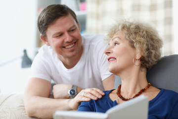 An elderly woman is sitting on sofa and smiling at young man. Relationship between adult children and parents concept