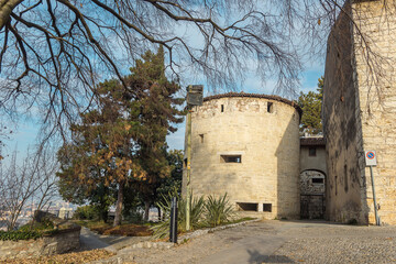 Fototapeta na wymiar Part of the castle of the city of Brescia on a sunny winter day. A viewthrough the trees. Castello di Brescia, Lombardy, Italy. Medieval castle with battlements, a tower, drawbridge and ramparts. 