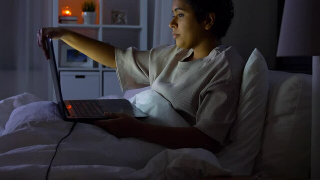 technology, internet, communication and people concept - young african american woman with laptop computer lying in bed at home at night