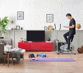 Man is doing sport and looking cell phone training program. Decorative living room, television unit, grey sofa purple mat and blue dumbbell brick wall home interior concept.