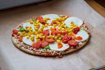 Colorful raw pizza before baking with mozzarella cheese, corn, pesto and cherry tomatoes. Frozen, unbaked, gluten free pizza placed on a baking sheet before putting it in the oven. Side view.