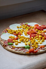 Colorful raw pizza before baking with mozzarella cheese, corn, pesto and cherry tomatoes. Frozen, unbaked, gluten free pizza placed on a baking sheet before putting it in the oven. Side view.