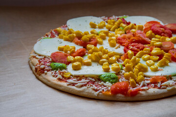 Colorful raw pizza before baking with mozzarella cheese, corn, pesto and cherry tomatoes. Frozen, unbaked, gluten free pizza placed on a baking sheet before putting it in the oven. Side view.
