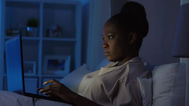 technology, internet, communication and people concept - young african american woman with laptop computer lying in bed at home at night