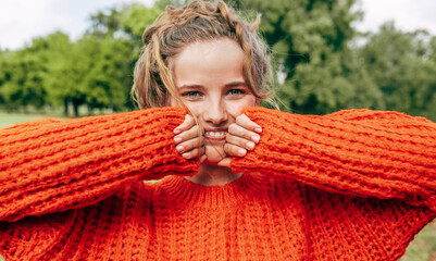 Closeup of a gorgeous young woman smiling in the park. Female wearing an orange sweater has joyful expression on the nature background.