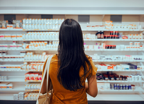 Rear View Of Young Woman With Bag Standing Against Shelf In Pharmacy Searching For Medicine