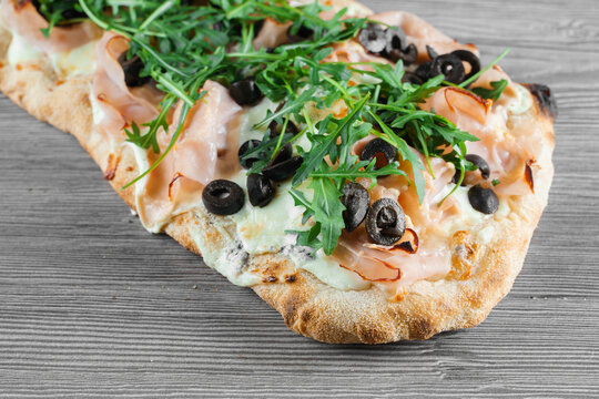 Pinsa romana gourmet italian cuisine on grey wooden background. Scrocchiarella traditional dish. Food delivery from pizzeria. Pinsa with meat, arugula, olives, cheese.