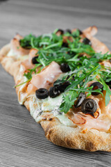 Pinsa romana gourmet italian cuisine on grey wooden background. Scrocchiarella traditional dish. Food delivery from pizzeria. Pinsa with meat, arugula, olives, cheese.