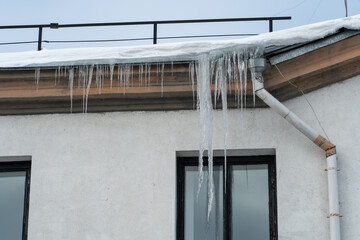 The roofs of the buildings are covered with snow and ice after a snowfall. Icicles hang from the facades of buildings. The fall of icicles carries a danger to people's lives. The frozen drainpipe