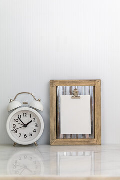 Wood frame clipboard with white notecard mockup with white alarm clock on marble portrait copy space