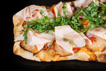 Pinsa romana gourmet italian cuisine on black wooden background. Scrocchiarella traditional dish. Food delivery from pizzeria. Pinsa with meat, arugula, olives, cheese.