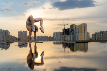 Flexible female gymnast doing handstand and calisthenic with reflection in the water on cityscape background during dramatic sunset