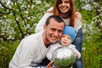 family, leisure and people concept - happy mother, father and little son with ball playing soccer at summer park. Portrait of mom, dad and little son on a background of flowering trees in the garden.