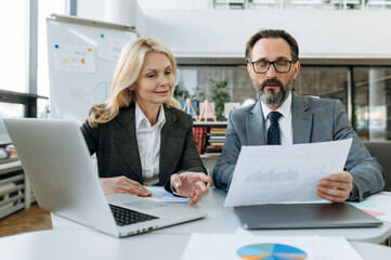 Concentrated mature business partners learning financial graphs for new project, doing paperwork. Elegant woman and serious man in formal suits sitting at the desk, look at the documents, teamwork