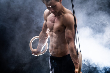 Muscular build man doing calisthenics on gymnastics rings indoor on black, smoked background. concept of motivation, desire and passion