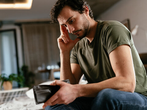 Young man checking empty wallet, no money. Unemployed sad man sitting at home showing empty wallet.