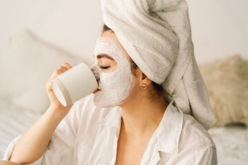 Beautiful woman applying facial mask and drinking tea in bed