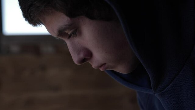 A closeup, profile shot of a sad looking, young teenage boy. The teen is sitting by himself in a garage, pondering and thinking with a sombre look on his face.
