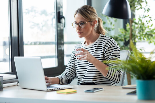 Beautiful Mature Business Woman Working With A Laptop While Drinking Glass Of Water On A Desk In The Office At Home.