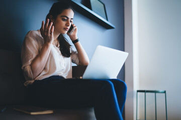 Confused young ethnic businesswoman speaking on smartphone and working remotely on laptop