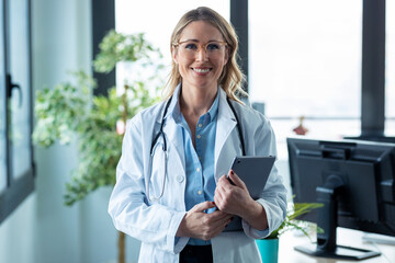 Attractive mature female doctor looking at camera while holding digital tablet standing in the consultation. - Powered by Adobe