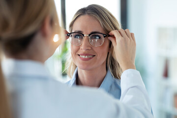 Young female doctor choosing while proving eyeglasses to mature beautiful patient in medical...