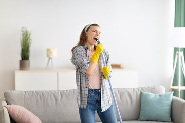 House cleaning is fun. Cheerful young lady using mop as mic, singing song while doing cleanup at...