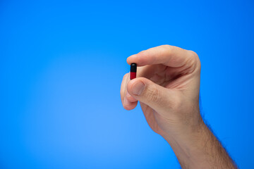 Red and black capsule medical pill held between fingers by Caucasian male hand isolate on blue background studio shot