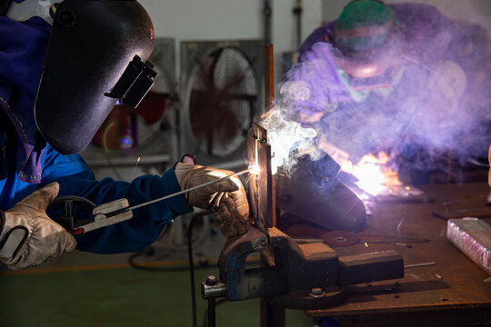 Two welder are assembling the workpiece by process shielded metal arc welding (SMAW). Welder in blue uniform, leather gloves, welding mask. The workers are testing a welding in the workshop - Powered by Adobe