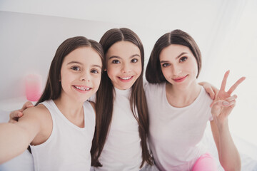Photo of sweet funny three siblings nightwear sitting bed smiling hugging tacking selfie showing v-sign indoors inside room home