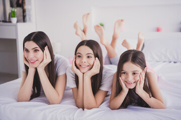 Photo of sweet adorable three sisters sleepwear lying bed having rest smiling arms cheekbones inside indoors home room