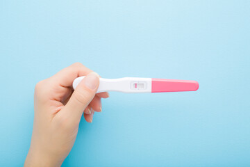 Young adult woman hand holding pregnancy test with one stripe on light blue table background....