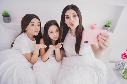 Photo of pretty cute three sisters sleepwear sitting bed having rest tacking selfie modern gadget sending kiss inside indoors home room - Powered by Adobe