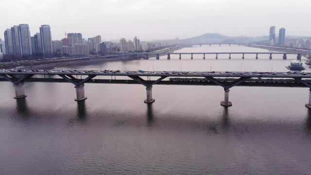 Cinematic aerial shot of Seoul and Han River, metro train ride at low deck of duplex bridge, camera fly to left, follow subway car motion. Congestion of motor traffic on top road